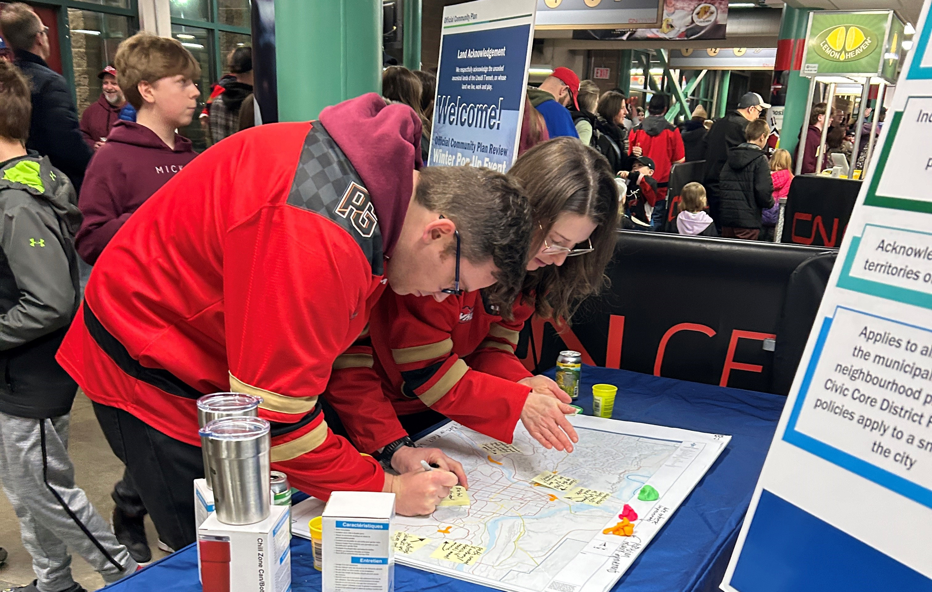 Two people writing on post-it notes, which are being added to a large map. large crowd in background.