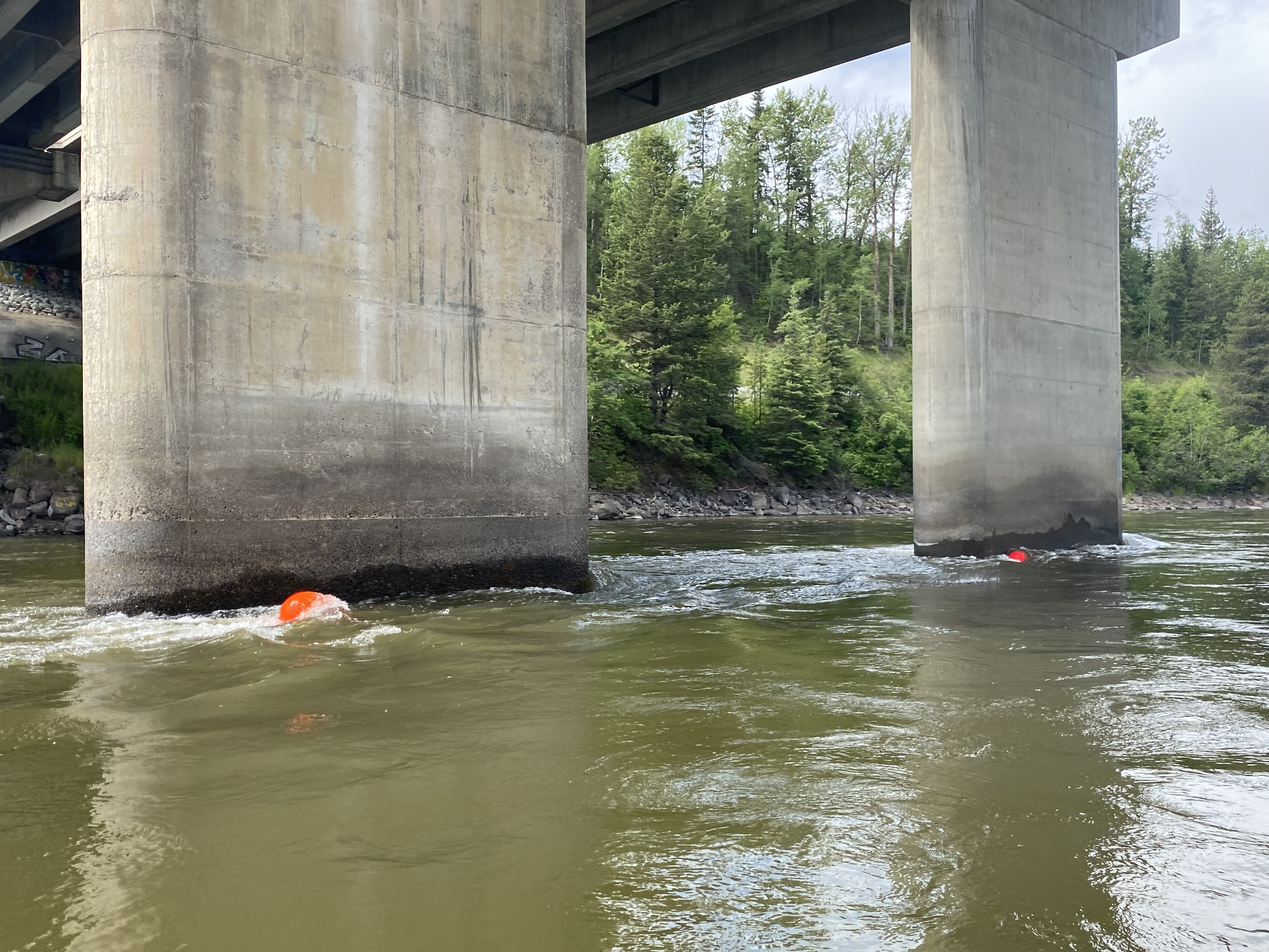 the section of river underneath a bridge showing two orange buoys floating in front of two concrete pillars