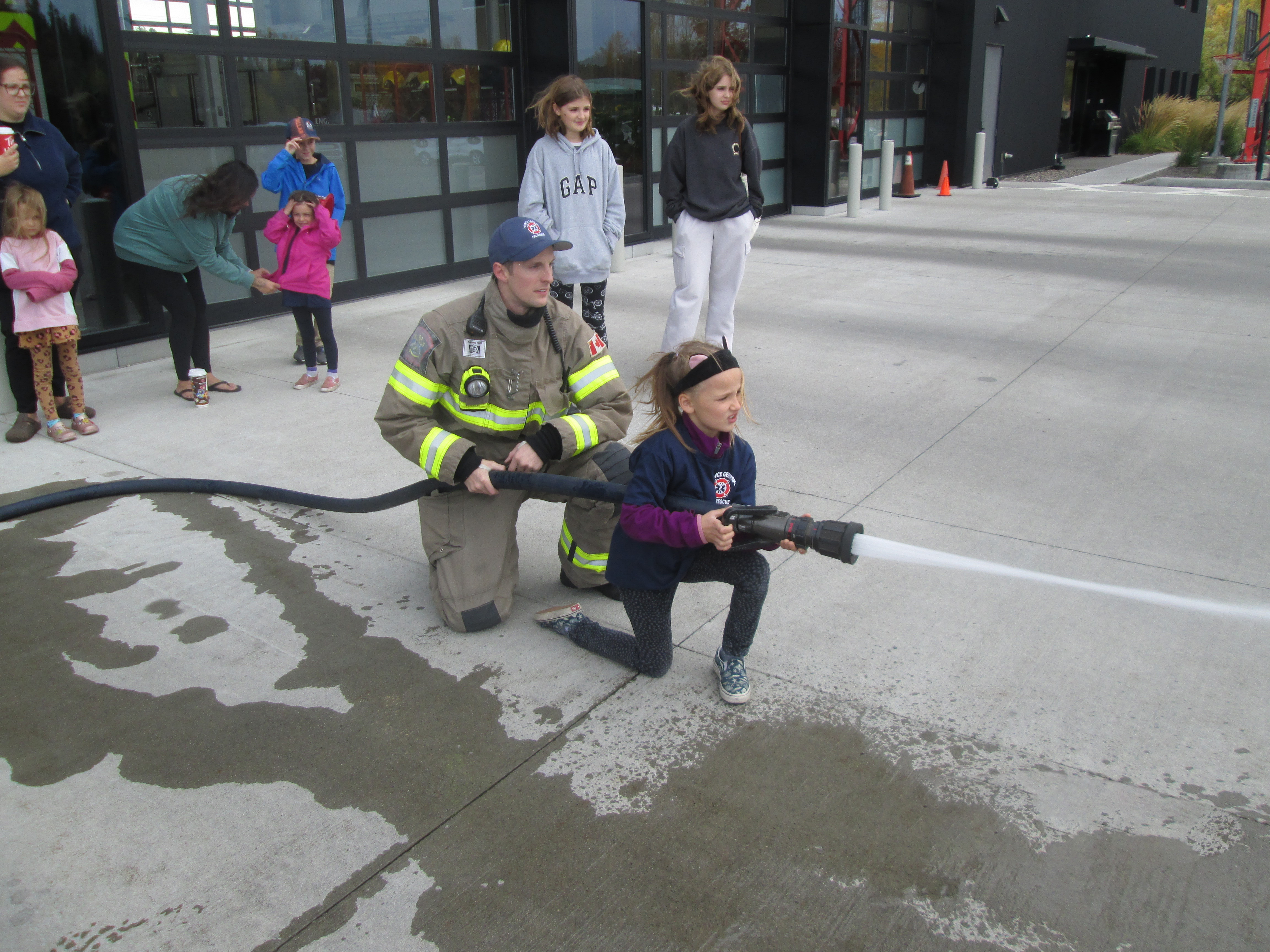 A young girl crouching down with a firefighter behind her both holding a large hose spraying water