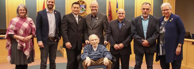 An older man (Trelle Morrow) surrounded by city councilors in Council Chambers.