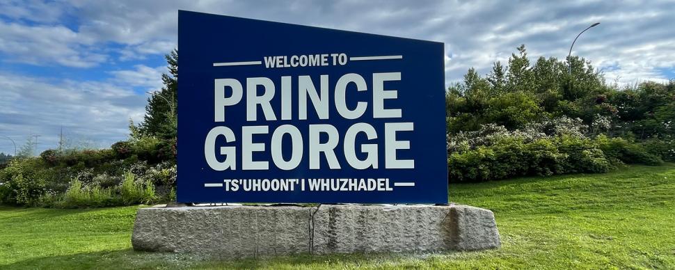 Blue sign on a stone base. White bold text on sign reads "WELCOME TO PRINCE GEORGE. TS'UHOONT'L WHUZHADEL"