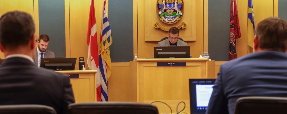 Two men seated at desk making a presentation in Council Chambers, with official flags and city crest in background