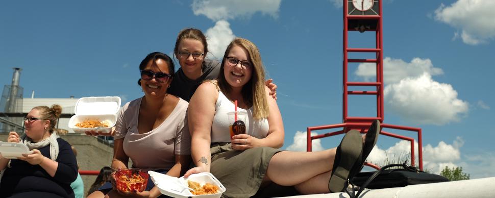 Photo of three people showing their food orders to the camera while sitting in front of a red clock tower, in Canada Games Plaza.
