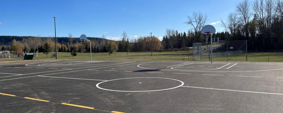 Outdoor basketball court with new blacktop and freshly painted lines. Leafless trees in background with bright blue sky.