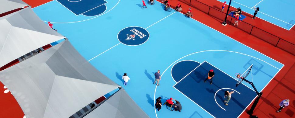 a blue paved basketball court showing some seating on the sidelines under big grey umbrellas