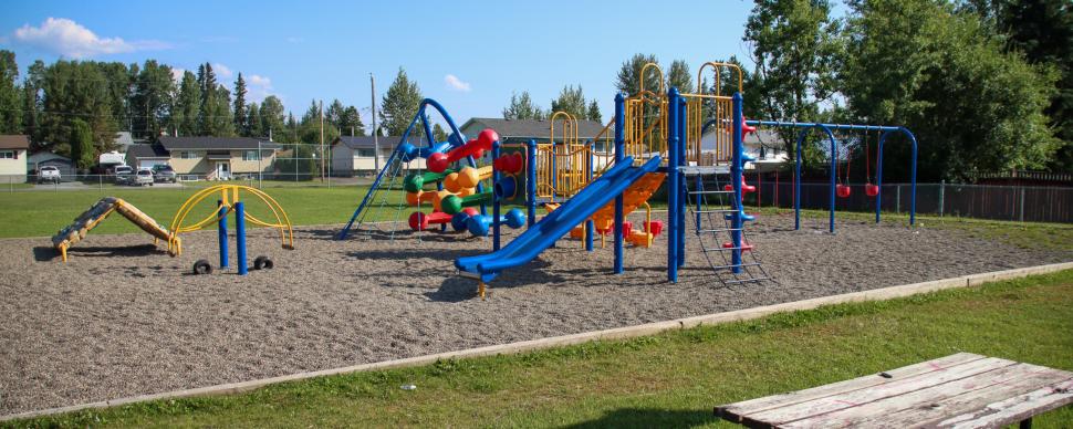 Colourful playground equipment with slide and climbing features surrounded by green grass on a sunny day.