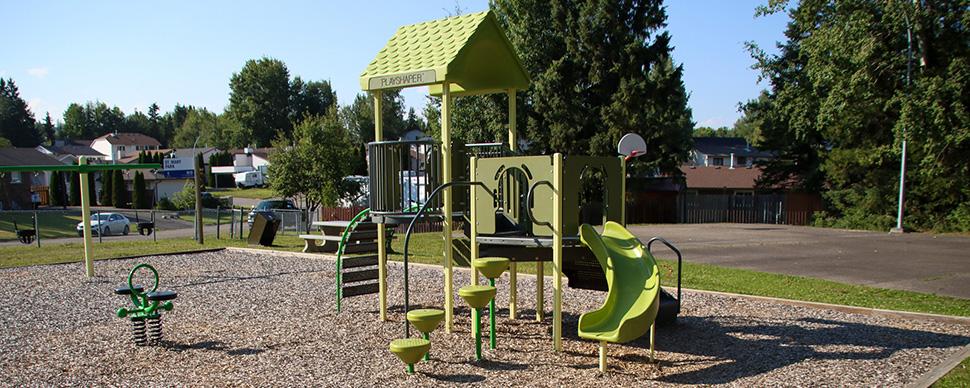 Green coloured playground equipment with slide on a sunny day. Picnic table and basketball court in background.