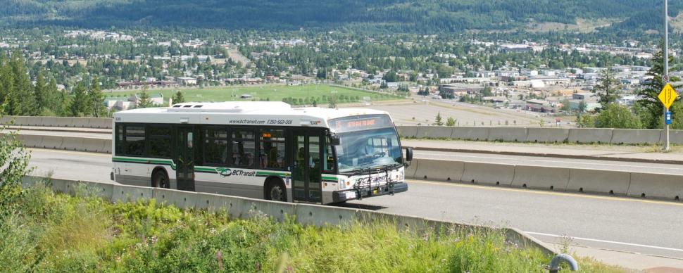 A city bus is travelling on a 4 lane road with a view of a city in the background.