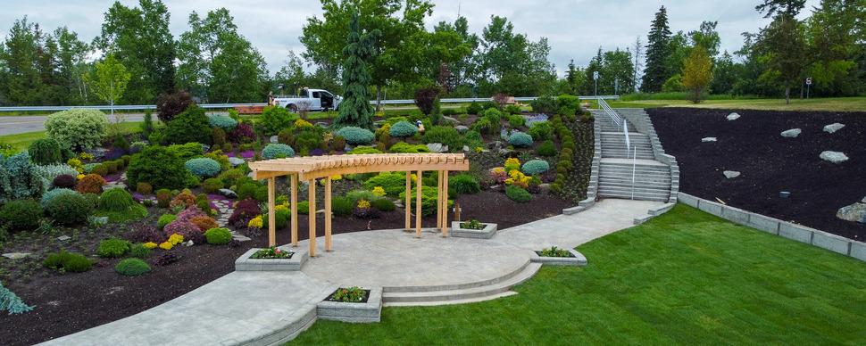 Park garden with wide cement pathway and staircase. A wooden structure stands on the cement path in front of the garden.