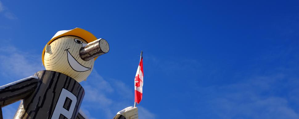 Mr. PG, the large mascot of Prince George, is standing outdoors on a clear blue-sky day, holding the Canadian flag
